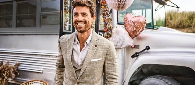 Lachender Mann der einen braunen Anzug trägt und vor einem Hochzeitsauto steht