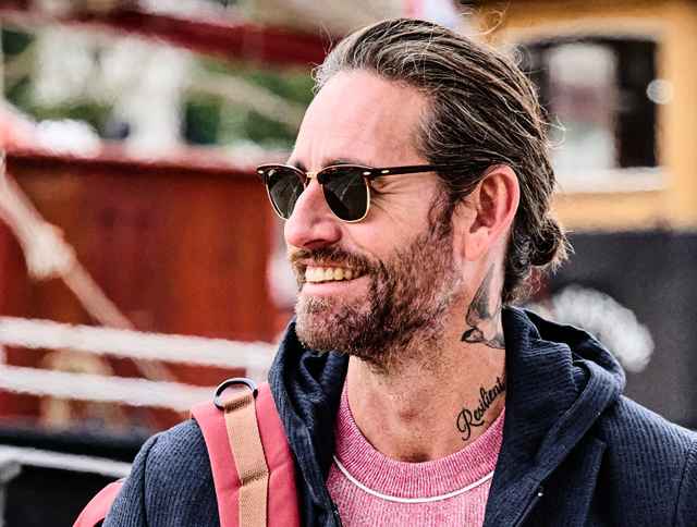 Ein Mann trägt eine dunkle Sonnenbrille, eine blaue Sweatshirtjacke und blickt lächelnd zur Seite.
