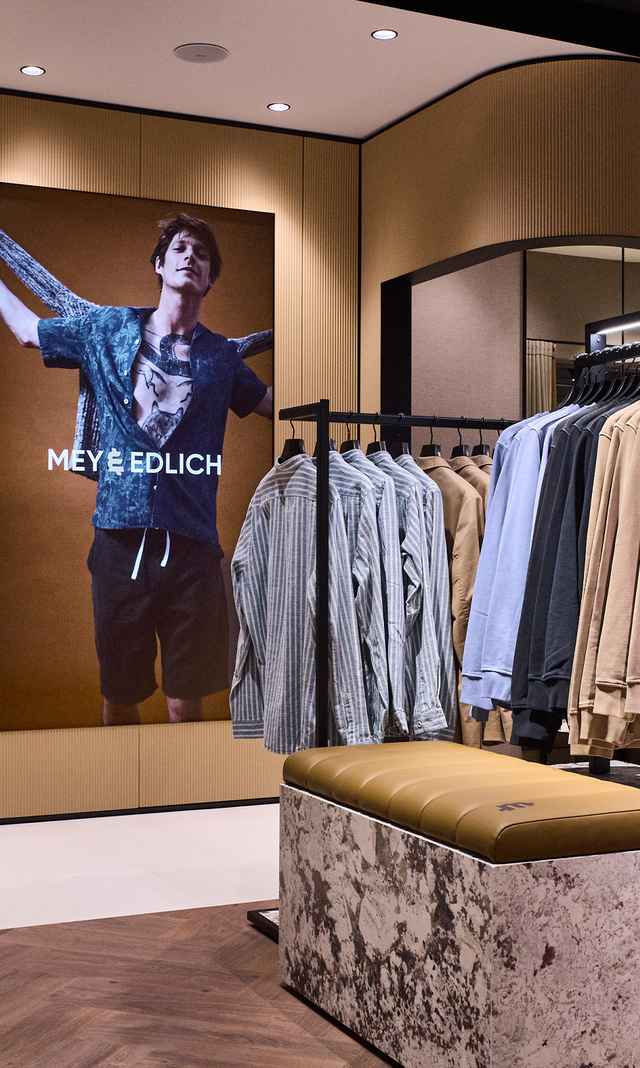 Das Bild zeigt die neu gestaltete M&E Filiale mit modernem Innenausbau, übersichtlich präsentierter Herrenbekleidung und einem großformatigen Markenmotiv an der Wand.