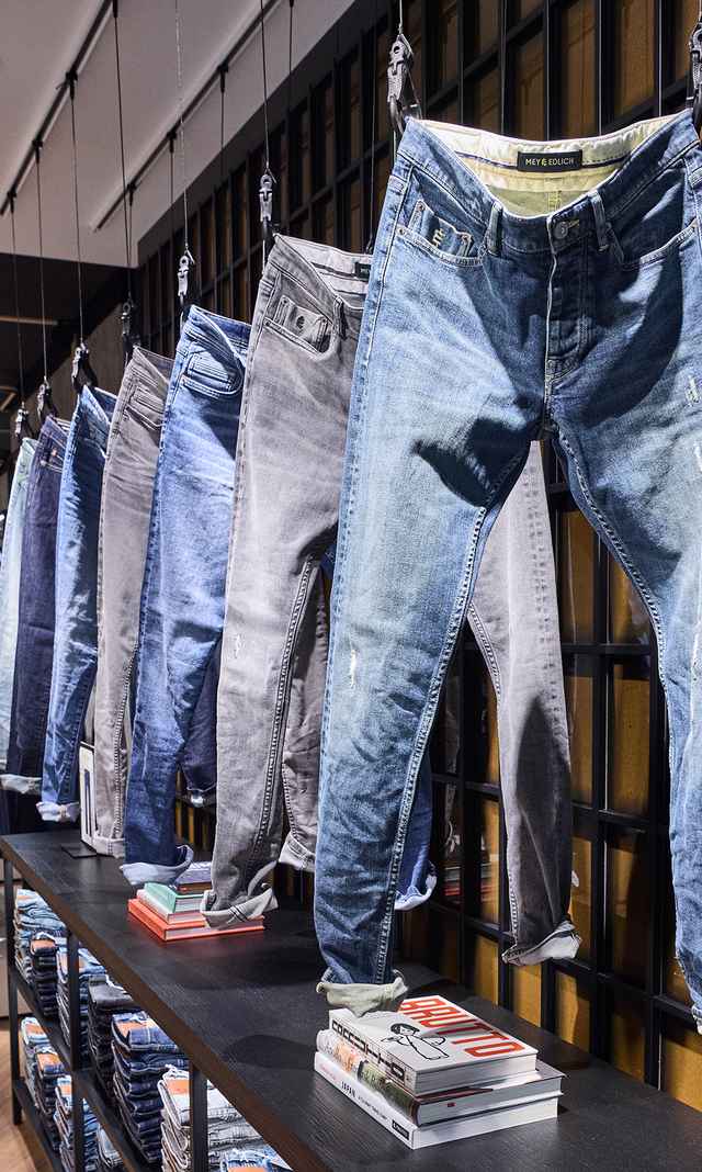 Das Bild zeigt eine modern inszenierte Jeanspräsentation in der neu gestalteten M&E Filiale mit mehreren aufgehängten Denim‑Hosen in unterschiedlichen Waschungen und Farben.