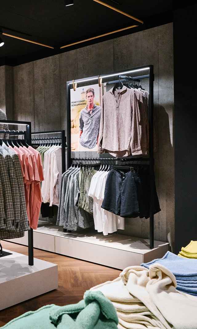 Das Bild zeigt die M&E Filiale mit einer modern beleuchteten Wandpräsentation aus Hemden, T‑Shirts und Shorts sowie einem integrierten Markenmotiv.
