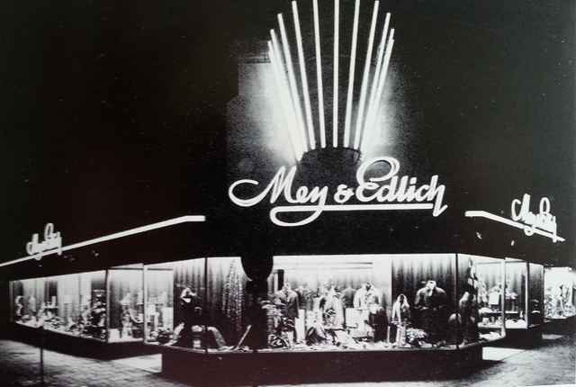 Schwarz-Weiß-Foto eines beleuchteten Schaufensters eines Modegeschäfts mit der Aufschrift "Mey & Edlich", dekoriert mit Kleidung und Accessoires.