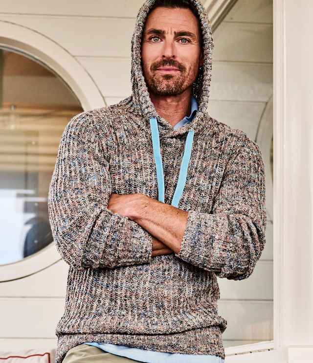 Ein Mann trägt einen grob gestrickten Strickpullover und steht mit verschränkten Armen vor einer Schiffswand.