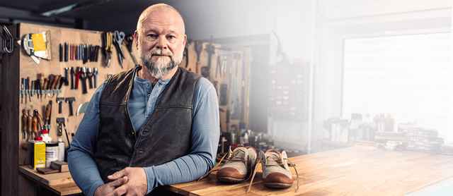 Ein älterer Mann in einer Werkstatt lehnt an einem Tisch mit Schuhen. Im Hintergrund sind Werkzeuge und Regale zu sehen.