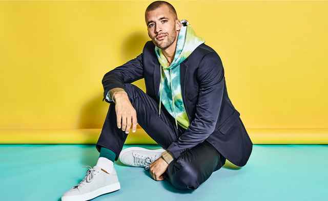 Sitzender Mann vor einem gelben Hintergrund in einem blauen Anzug und bunten Hoodie