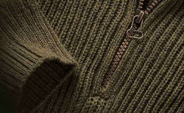 Das Bild zeigt eine Nahaufnahme eines grob gestrickten Merinowollpullovers in Olivgrün mit einem metallenen Reißverschluss.