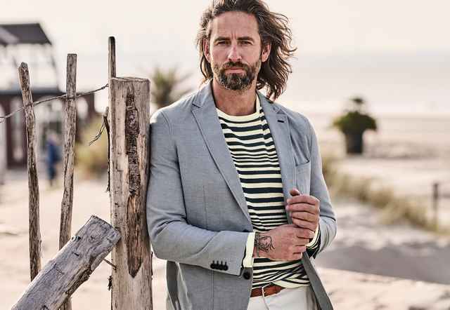 Mann mit langem Haar und Bart posiert in gestreiftem Hemd und grauem Blazer vor einer Strandkulisse mit Holzzaun und Sand.