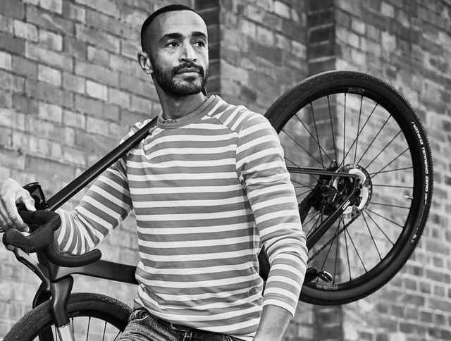 Eine schwarz-weiß Optik eines Mannes in einem gestreiften Langarmshirt trägt ein Fahrrad über seiner Schulter.