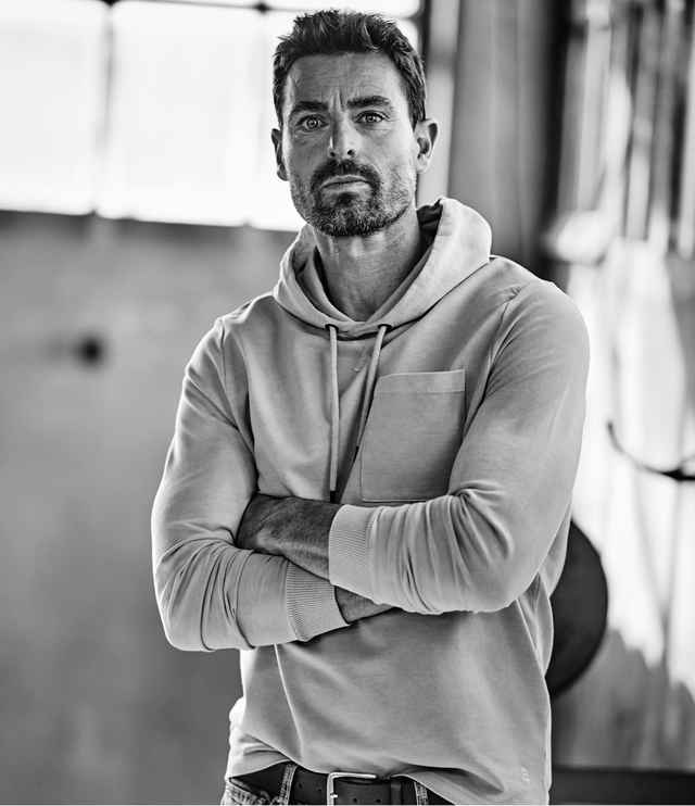 Ein schwarz-weiß Bild von einem Mann der mit einem Hoodie bekleidet und mit verschränkten Armen in einer Sporthalle steht.