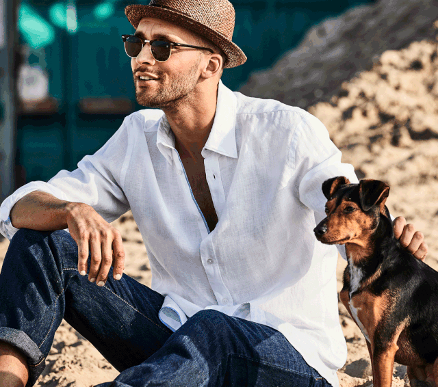Mann mit Hut, Sonnenbrille und weißen Leinenhemd sitzt auf einem Fels neben einem kleinen Hund.