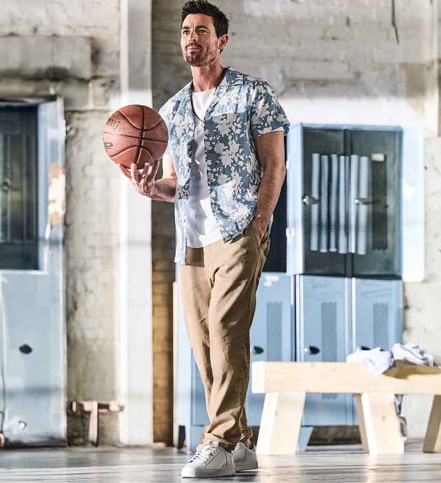 Ein Mann in lässiger Kleidung hält einen Basketball in einer industriellen Umgebung, mit Spinden im Hintergrund.