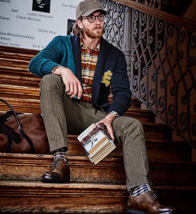 Mann mit Brille und Kappe sitzt auf einer Treppe, hält Bücher und trägt kariertes Hemd, Strickjacke und Tweed-Hose. Große Tasche neben ihm.