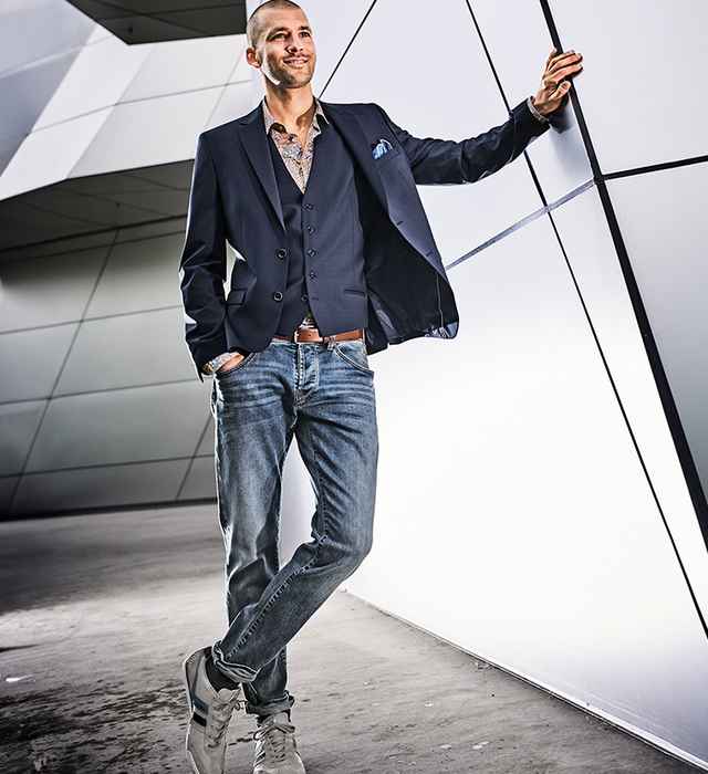 Mann in dunklem Anzug und Jeans lehnt lässig an einer futuristischen Wand. Er lächelt und trägt sportliche Schuhe.