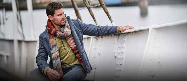 Ein Mann trägt eine olivgrüne Cord-Hemdjacke, darüber ein graues Sakko und einen bunten, gestrickten Schal in Herbstfarben