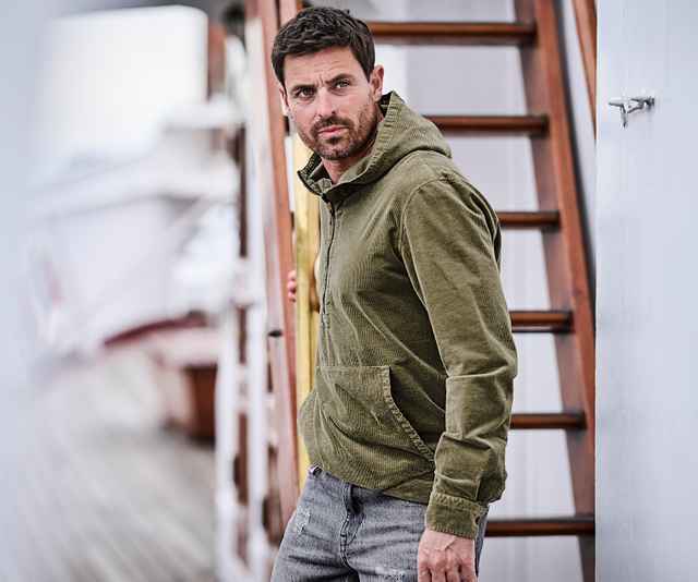 Ein Mann trägt eine olivgrüne Cordjacke mit Kapuze und Bauchtasche, kombiniert mit einer grauen Jeans.