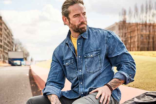 Mann mit Bart und Tattoos sitzt in einem Jeanshemd auf einer Mauer, blickt nach links. Im Hintergrund städtische Straße und Gebäude.