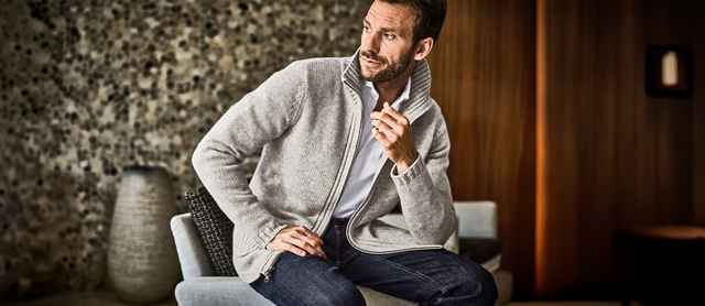 Ein Mann lehnt entspannt an einem Stuhl und trägt eine hellgraue Strickjacke.