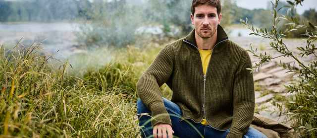 Ein Mann sitzt im Grünen und trägt eine olivgrüne Strickjacke mit Reißverschluss über einem gelben T-Shirt und einer blauen Jeans.