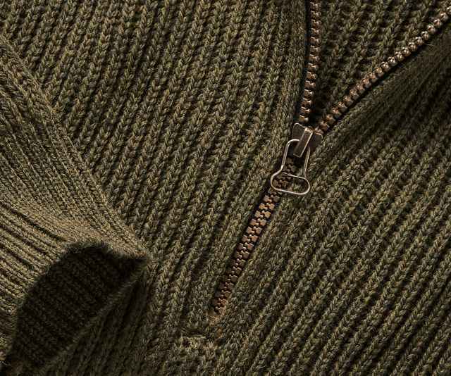 Das Bild zeigt eine Nahaufnahme einer grob gestrickten, olivgrünen Strickjacke mit einem metallischen Reißverschluss.