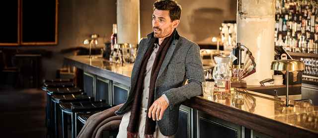 Ein Mann lehnt in einer stilvollen Bar an der Theke, trägt einen grauen Blazer und einen Schal, während vor ihm ein Glas und eine Whiskyflasche stehen.