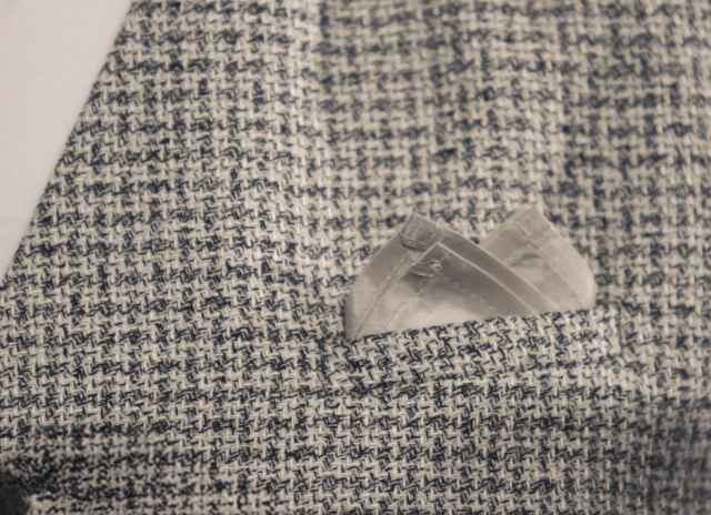 Nahaufnahme eines Tweed-Sakkos mit grau-weißem Muster, das ein ordentlich gefaltetes Einstecktuch in der Brusttasche trägt.