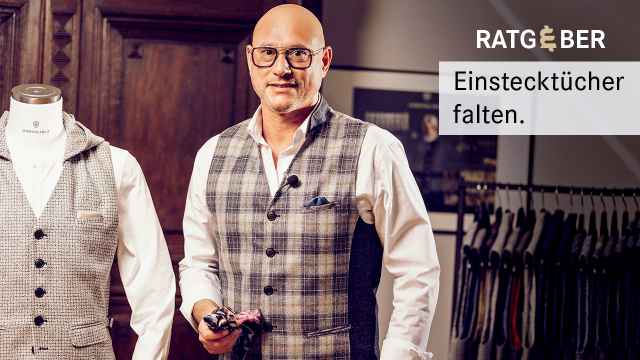 Ein Mann in Anzugweste faltet Einstecktücher neben einer Schaufensterpuppe in einem Ankleideraum.