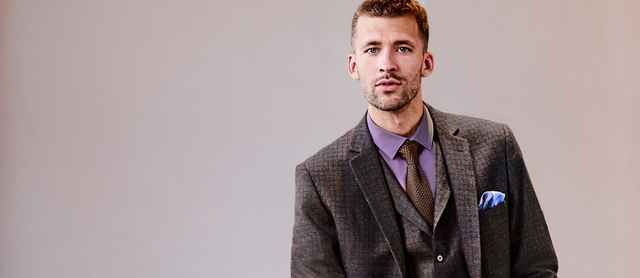 Ein Mann in einem eleganten, dreiteiligen Anzug mit violettem Hemd und blauer Einstecktuch steht vor neutralem Hintergrund.