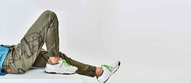 Person trägt olivgrüne Hose und weiße Turnschuhe mit grünen Details, Beine entspannt auf einer weißen Oberfläche ausgestreckt.