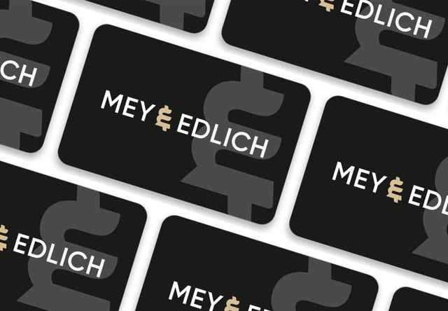 Auf dem Bild sind mehrere schwarze Karten mit dem weißen „MEY & EDLICH“-Logo und einem großen grauen grafischen Element im Hintergrund diagonal angeordnet.