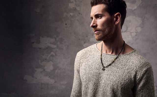 Eine Person trägt einen hellen Strickpullover und eine Halskette und steht vor einer grauen, strukturierten Wand.