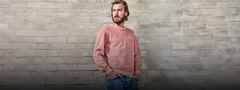 Ein Mann mit Bart trägt einen rosa Pullover und blaue Jeans, steht vor einer grauen Ziegelwand und blickt nach rechts.
