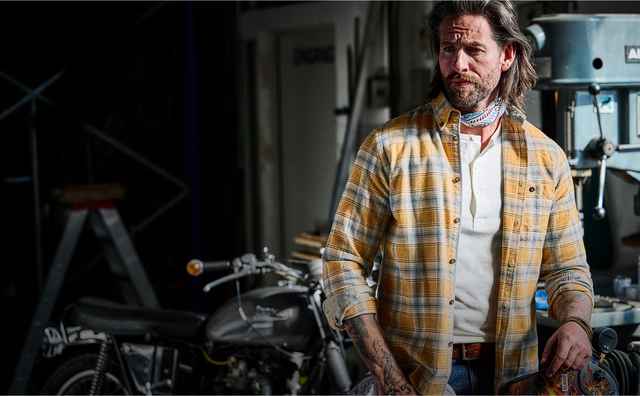 Mann mit Bart und kariertem Hemd steht in einer Werkstatt neben einer Maschine und einem Motorrad, mit einer entschlossenen Miene.