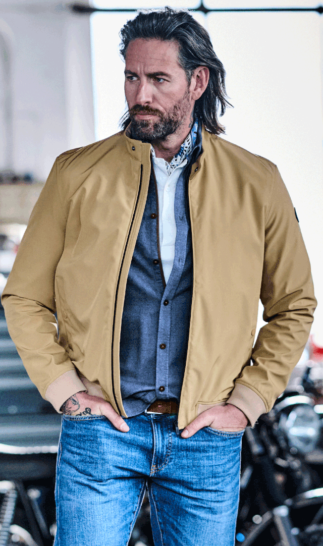 Ein Mann mit Bart trägt eine beige Jacke, Jeans und ein Hemd. Er steht in einer Garage vor einem unscharfen Hintergrund mit Motorrädern.