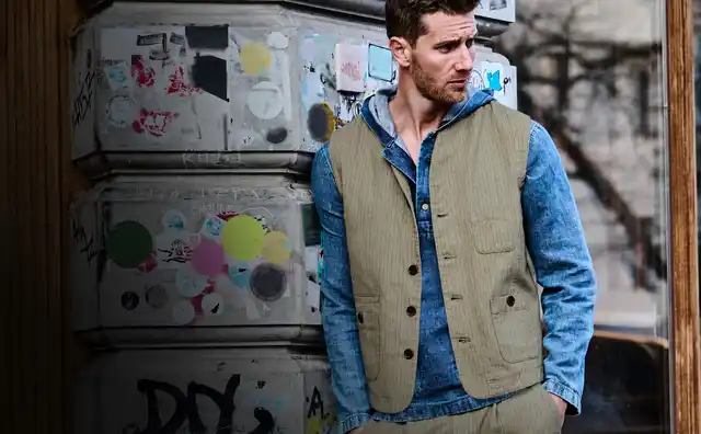 Ein Mann in blauer Jeansbluse und beiger Weste lehnt an einer mit Graffiti bedeckten Wand in einer urbanen Umgebung.