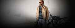 Mann mit Sonnenbrille und Bart steht neben einem Motorrad. Er trägt eine beige Jacke über einem dunkel gestreiften Pullover und hält eine Tasche.