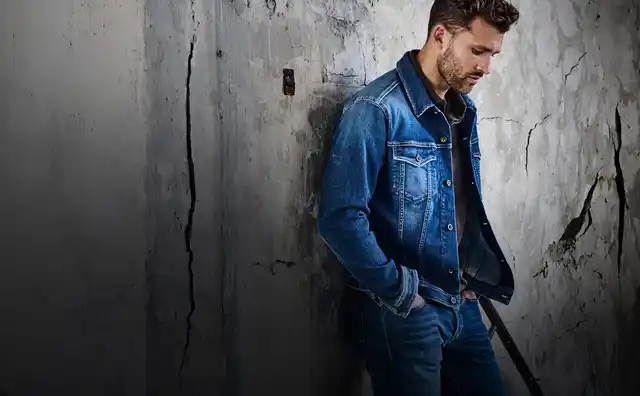 Mann in Jeansjacke und Jeans steht nachdenklich gegen eine alte, rissige Betonwand gelehnt.