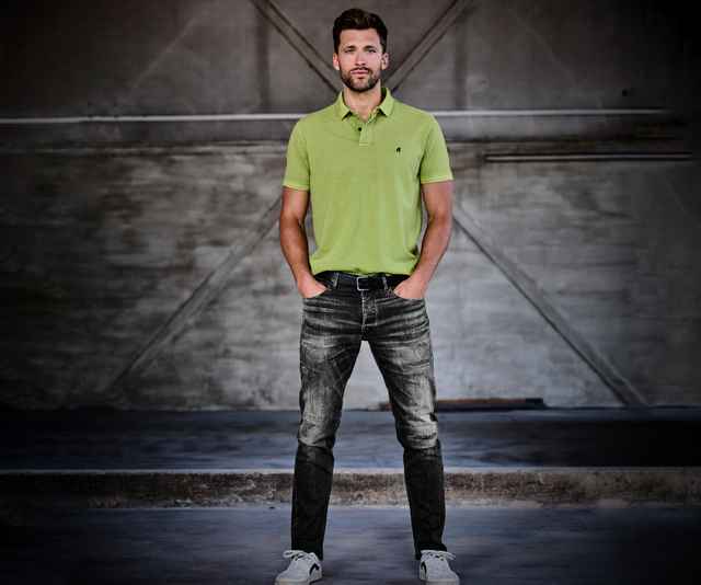 Mann in grünem Poloshirt und Jeans steht mit Händen in den Taschen vor einer grauen, industriellen Wand.