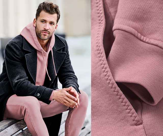 Mann sitzt auf einer Bank, trägt einen rosa Hoodie und Hose mit schwarzem Mantel. Nahaufnahme des rosa Stoffes zeigt Details der Textur.
