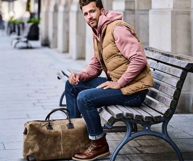Ein Mann in rosa Kapuzenpullover und beiger Weste sitzt auf einer Bank neben einer braunen Reisetasche auf einer gepflasterten Straße.