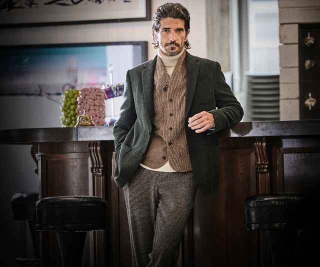 Ein Mann in eleganter Kleidung steht lässig an einer Bar, hinter ihm sind Trauben und Dekoration.