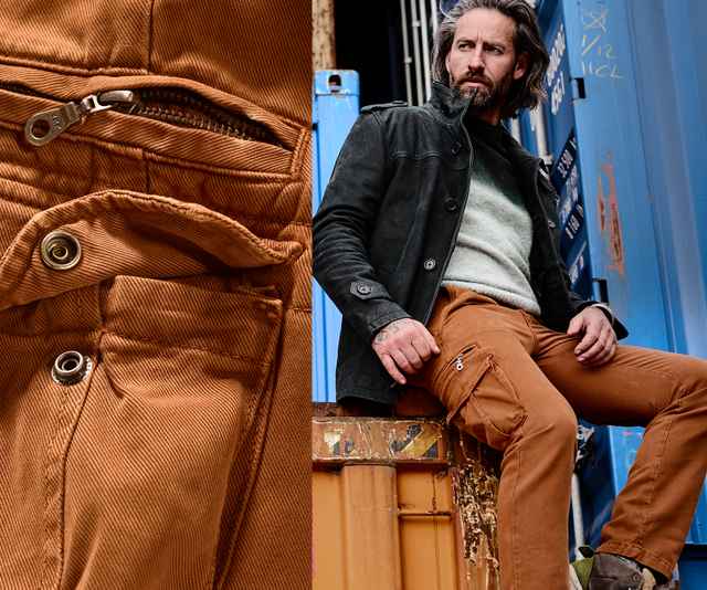 Mann in schwarzer Jacke und brauner Hose sitzt auf einem Container; Detail eines Reißverschlusses und Knopfes der Hose.