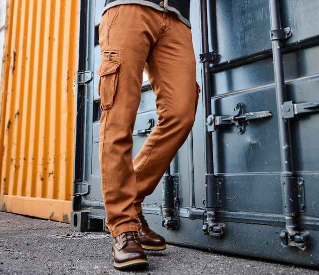 Person trägt orangebraune Cargo-Hose und braune Stiefel, steht vor einem blauen und gelben Schiffscontainer auf Asphalt.
