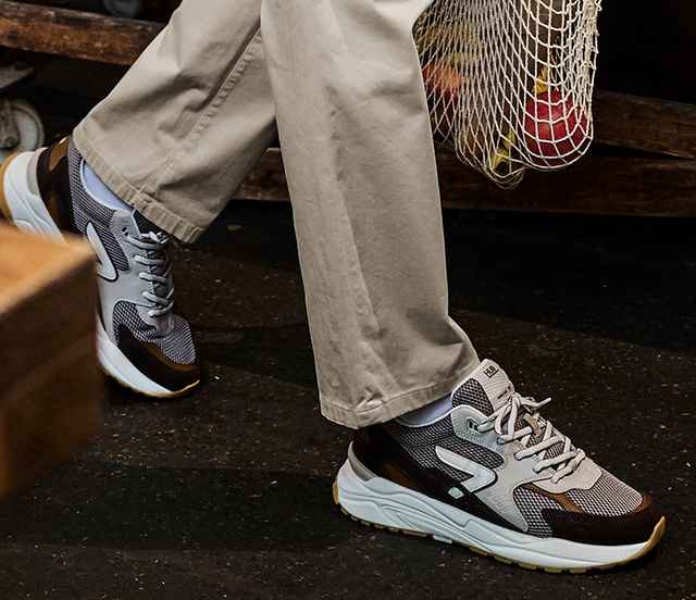 Person mit beigen Hosen trägt graue Sneaker und hält ein Netz mit Obst.