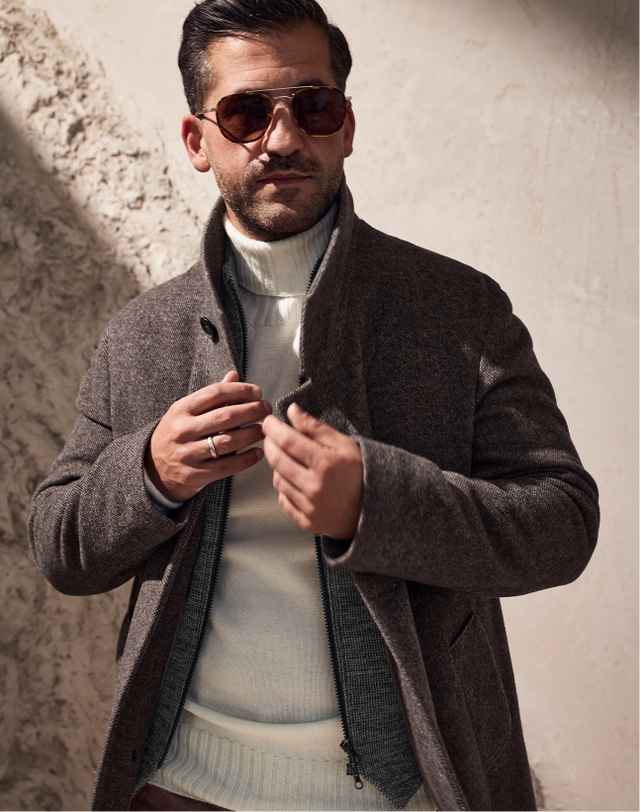 Mann in braunem Mantel und weißem Rollkragenpullover, trägt Sonnenbrille, steht vor einer strukturierten Wand.