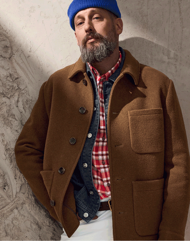 Ein Mann mit Bart trägt eine braune Jacke über einem karierten Hemd und einer Jeansjacke. Er hat eine blaue Mütze auf.