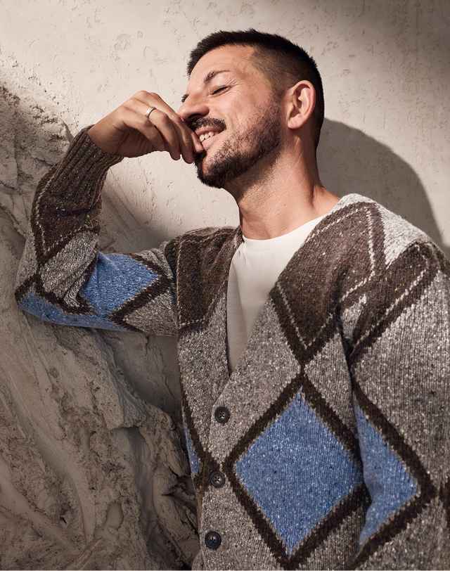 Mann trägt eine gestrickte Strickjacke mit Rautenmuster in Braun, Grau und Blau, steht lächelnd und mit erhobener Hand an einer Steinmauer.