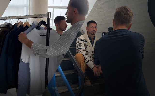 Männer im Fotostudio; links wird fotografiert, in der Mitte hängen Hemden auf Ständer, rechts sitzen zwei Männer in Gespräch.