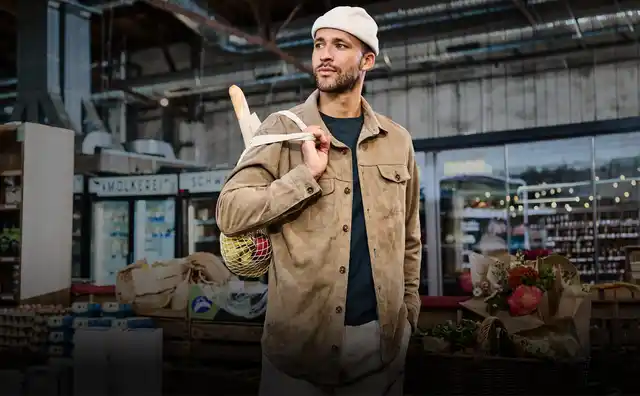 Ein Mann in einem beige Outfit steht mit einer Einkaufstasche voller Gemüse und einem Baguette in einem gut beleuchteten Supermarkt.