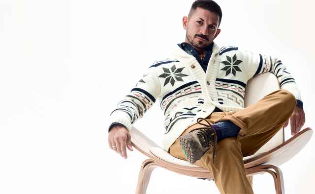 Mann in weißem, gemustertem Cardigan und beige Hose sitzt lässig auf einem modernen Stuhl vor hellem Hintergrund.