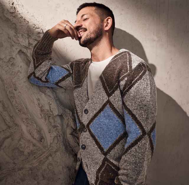 Mann in gemustertem Cardigan lehnt lächelnd an einer strukturierten Wand. Natürliche Licht- und Schatteneffekte heben Details hervor.